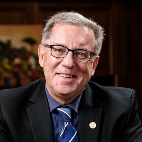 Portrait of Prof. T. Eugene Cloete, seated at a wooden desk in an office, wearing a dark suit and blue striped tie